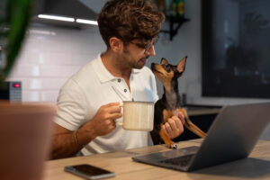 Homem de óculos e camisa branca segurando uma xícara enquanto olha para um pequeno cachorro preto e marrom, que está em seu colo. Ele está sentado em uma cozinha moderna, com um laptop aberto à sua frente e um celular sobre a mesa