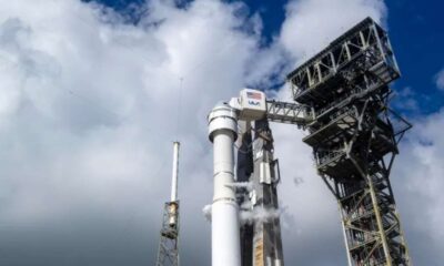 A imagem mostra a espaçonave Starliner, da Boeing, acoplada ao foguete Atlas V em uma plataforma de lançamento metálica, com nuvens no céu ao fundo. A estrutura de suporte ao lado do foguete exibe o logotipo da ULA (United Launch Alliance).