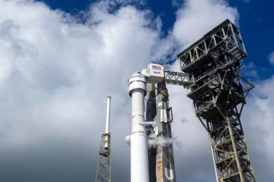 A imagem mostra a espaçonave Starliner, da Boeing, acoplada ao foguete Atlas V em uma plataforma de lançamento metálica, com nuvens no céu ao fundo. A estrutura de suporte ao lado do foguete exibe o logotipo da ULA (United Launch Alliance).