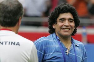 Maradona veste camisa azul listrada e colar com cruz, enquanto conversa com outra pessoa vestindo uma camisa branca com a palavra 'Argentina' escrita nas costas.