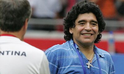 Maradona veste camisa azul listrada e colar com cruz, enquanto conversa com outra pessoa vestindo uma camisa branca com a palavra 'Argentina' escrita nas costas.