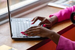 Mãos de uma mulher digitando em um laptop, usando unhas longas e rosa, com uma blusa rosa vibrante.