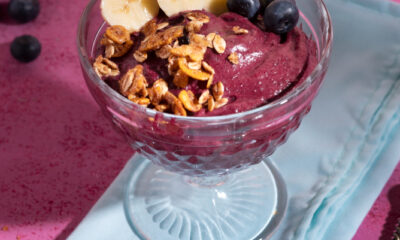Taça de vidro com açaí cremoso decorado com fatias de banana, mirtilos e granola crocante, sobre guardanapo azul em superfície rosa; ao fundo, pedaços de limão e tigela com mirtilos.