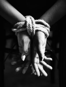 Mãos de uma pessoa amarradas com corda, representando situação de restrição ou violência. A imagem está em preto e branco, com foco nos detalhes da amarração e das mãos entrelaçadas.