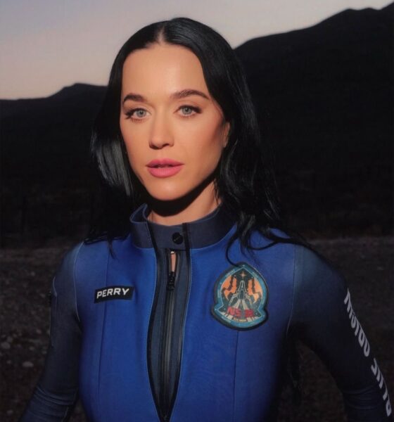 Katy Perry vestindo um macacão espacial azul da Blue Origin, com seu nome e o emblema da missão estampados no uniforme, posando em um cenário ao ar livre com montanhas ao fundo, iluminada pela luz do pôr do sol.