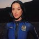 Katy Perry vestindo um macacão espacial azul da Blue Origin, com seu nome e o emblema da missão estampados no uniforme, posando em um cenário ao ar livre com montanhas ao fundo, iluminada pela luz do pôr do sol.