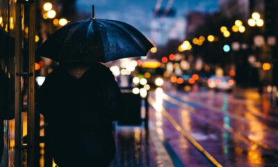 Pessoa caminha sob chuva à noite segurando um guarda-chuva preto em uma calçada iluminada por luzes urbanas e faróis de carros.