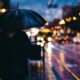 Pessoa caminha sob chuva à noite segurando um guarda-chuva preto em uma calçada iluminada por luzes urbanas e faróis de carros.