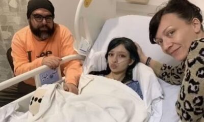 Adolescente deitada em cama hospitalar com sonda nasal, acompanhada por um homem com blusa laranja e uma mulher com blusa estampada, dentro de um quarto de hospital.