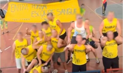 Grupo de estudantes vestindo camisetas amarelas posa para foto em quadra esportiva, com alguns participantes sobre os ombros de colegas. Ao fundo, faixa amarela exibe frase ofensiva com conotação violenta. Rosto dos envolvidos aparece desfocado. Cena está relacionada a caso de apologia ao estupro em evento universitário.