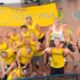 Grupo de estudantes vestindo camisetas amarelas posa para foto em quadra esportiva, com alguns participantes sobre os ombros de colegas. Ao fundo, faixa amarela exibe frase ofensiva com conotação violenta. Rosto dos envolvidos aparece desfocado. Cena está relacionada a caso de apologia ao estupro em evento universitário.