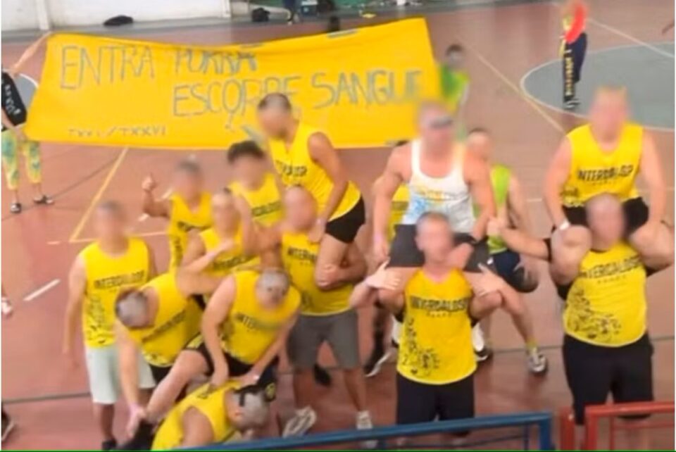 Grupo de estudantes vestindo camisetas amarelas posa para foto em quadra esportiva, com alguns participantes sobre os ombros de colegas. Ao fundo, faixa amarela exibe frase ofensiva com conotação violenta. Rosto dos envolvidos aparece desfocado. Cena está relacionada a caso de apologia ao estupro em evento universitário.