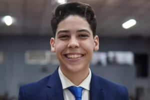 Adolescente de terno azul-marinho e gravata azul sorrindo, com fundo desfocado de ambiente interno, possivelmente uma igreja.