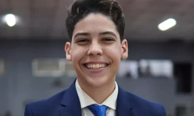 Adolescente de terno azul-marinho e gravata azul sorrindo, com fundo desfocado de ambiente interno, possivelmente uma igreja.