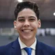 Adolescente de terno azul-marinho e gravata azul sorrindo, com fundo desfocado de ambiente interno, possivelmente uma igreja.