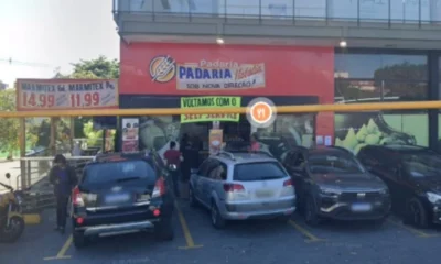 Fachada da padaria Natália, no bairro Serrano, em Belo Horizonte, com letreiro visível e vários carros estacionados em frente ao estabelecimento. Placa anuncia preços de marmitex.