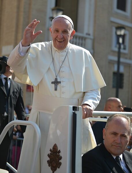 Papa Francisco sorri e acena para a multidão enquanto está em pé no papamóvel, usando sua tradicional batina branca, durante um evento ao ar livre no Vaticano.