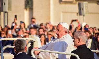 Papa Francisco aparece sentado em um veículo papal ao ar livre, vestindo batina branca e solidéu. Ao seu lado, um religioso de batina preta. Ao fundo, uma multidão fotografa e observa a cena, evidenciando um momento público e solene no Vaticano, com clima de respeito e atenção.