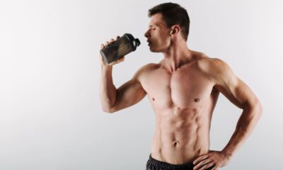 Homem jovem e musculoso, sem camisa, bebendo suplemento em um copo shaker, posicionado de perfil contra fundo claro