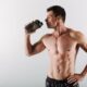 Homem jovem e musculoso, sem camisa, bebendo suplemento em um copo shaker, posicionado de perfil contra fundo claro
