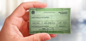 Pessoa segurando um título de eleitor verde da República Federativa do Brasil, com identificação biométrica, em fundo desfocado.