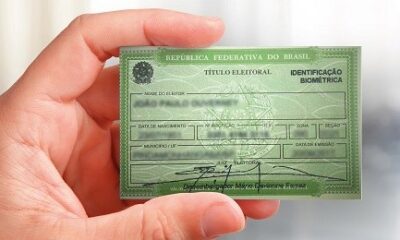 Pessoa segurando um título de eleitor verde da República Federativa do Brasil, com identificação biométrica, em fundo desfocado.