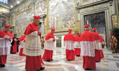 Cardeais da Igreja Católica caminham pela Capela Sistina durante cerimônia do conclave, vestindo trajes vermelhos tradicionais, sob afrescos renascentistas e vigilância da Guarda Suíça.