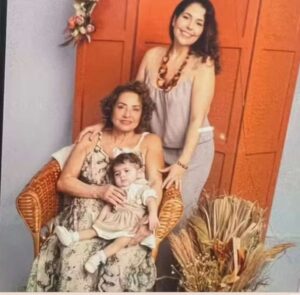 Três mulheres de uma mesma família posam para uma foto em ambiente interno. Uma mulher idosa está sentada em uma cadeira de vime segurando uma bebê no colo; atrás delas, uma mulher adulta, de pé, sorri. Ao fundo, uma porta de madeira laranja e um arranjo de flores secas decoram o espaço.
