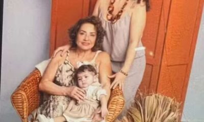 Três mulheres de uma mesma família posam para uma foto em ambiente interno. Uma mulher idosa está sentada em uma cadeira de vime segurando uma bebê no colo; atrás delas, uma mulher adulta, de pé, sorri. Ao fundo, uma porta de madeira laranja e um arranjo de flores secas decoram o espaço.