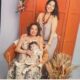 Três mulheres de uma mesma família posam para uma foto em ambiente interno. Uma mulher idosa está sentada em uma cadeira de vime segurando uma bebê no colo; atrás delas, uma mulher adulta, de pé, sorri. Ao fundo, uma porta de madeira laranja e um arranjo de flores secas decoram o espaço.