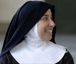 Freira sorri enquanto olha para o lado; ela usa hábito religioso preto com véu escuro e touca branca, em ambiente interno com fundo desfocado.