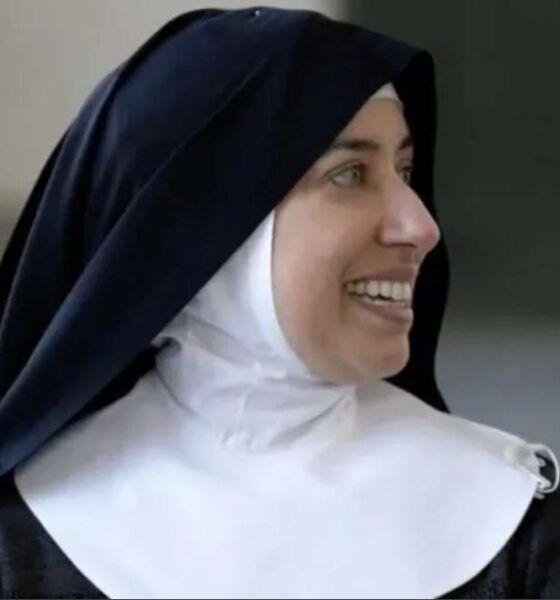 Freira sorri enquanto olha para o lado; ela usa hábito religioso preto com véu escuro e touca branca, em ambiente interno com fundo desfocado.