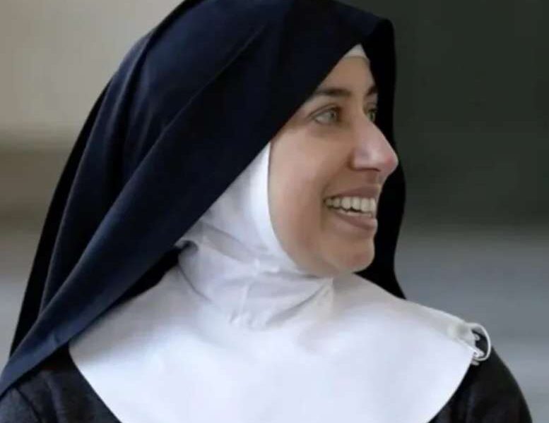 Freira sorri enquanto olha para o lado; ela usa hábito religioso preto com véu escuro e touca branca, em ambiente interno com fundo desfocado.