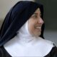 Freira sorri enquanto olha para o lado; ela usa hábito religioso preto com véu escuro e touca branca, em ambiente interno com fundo desfocado.