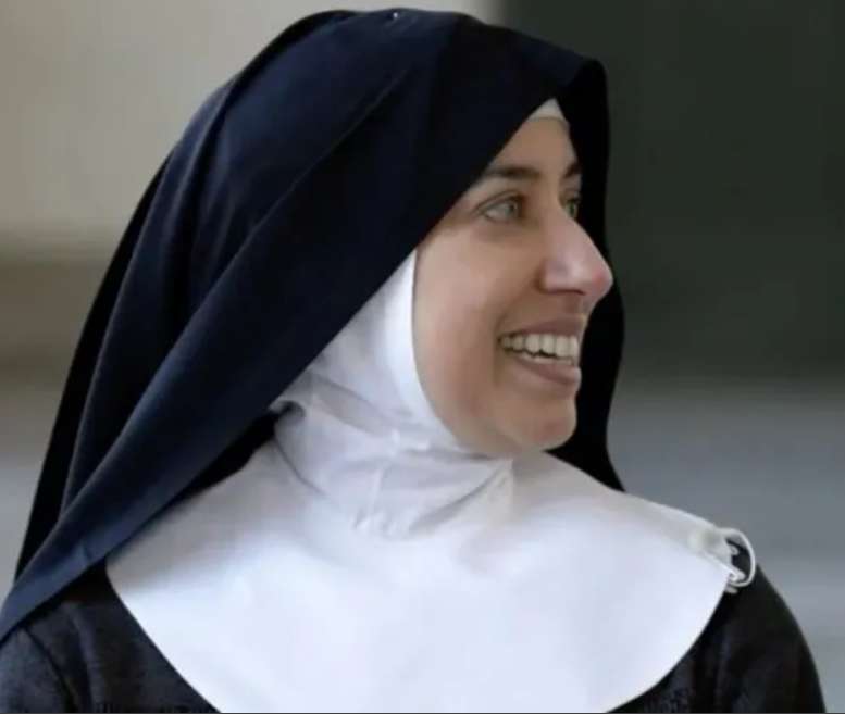 Freira sorri enquanto olha para o lado; ela usa hábito religioso preto com véu escuro e touca branca, em ambiente interno com fundo desfocado.