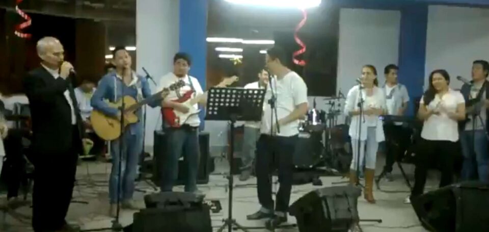 Papa Leão XIV, então bispo Roberto Prevost, canta ao microfone acompanhado por uma banda de jovens músicos durante uma celebração natalina; ao fundo, instrumentos musicais e decoração com fitas vermelhas indicam o clima festivo.