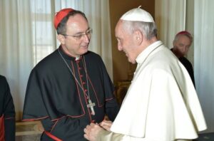 Dom Sergio da Rocha, vestido com traje e solidéu vermelho de cardeal, cumprimenta o papa Francisco, que usa veste branca e solidéu branco; ambos sorriem e trocam um aperto de mãos em ambiente interno bem iluminado, com cortinas claras ao fundo.