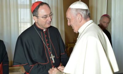 Dom Sergio da Rocha, vestido com traje e solidéu vermelho de cardeal, cumprimenta o papa Francisco, que usa veste branca e solidéu branco; ambos sorriem e trocam um aperto de mãos em ambiente interno bem iluminado, com cortinas claras ao fundo.