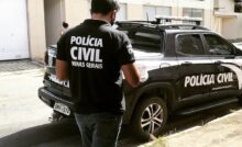Agente da Polícia Civil de Minas Gerais em pé próximo uma viatura oficial durante operação em área urbana, com prédios ao fundo.