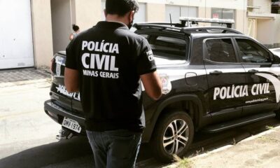 Agente da Polícia Civil de Minas Gerais em pé próximo a uma viatura oficial durante operação em área urbana, com prédios ao fundo.