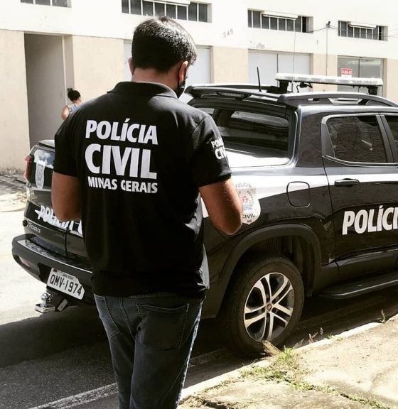 Agente da Polícia Civil de Minas Gerais em pé próximo a uma viatura oficial durante operação em área urbana, com prédios ao fundo.