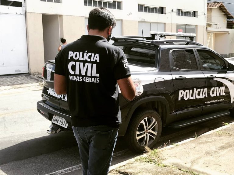 Agente da Polícia Civil de Minas Gerais em pé próximo a uma viatura oficial durante operação em área urbana, com prédios ao fundo.
