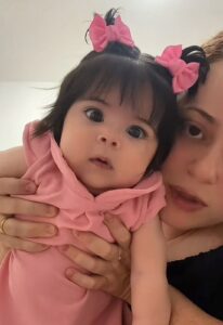 Mulher segura uma bebê de olhos grandes e cabelo escuro preso com dois laços rosas. A criança veste um vestido rosa e encara a câmera com expressão curiosa. A mãe aparece parcialmente ao lado, olhando na mesma direção.