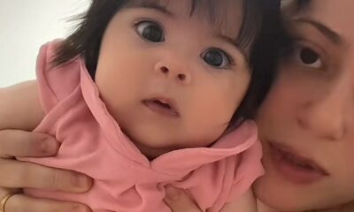 Mulher segura uma bebê de olhos grandes e cabelo escuro preso com dois laços rosas. A criança veste um vestido rosa e encara a câmera com expressão curiosa. A mãe aparece parcialmente ao lado, olhando na mesma direção.
