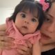 Mulher segura uma bebê de olhos grandes e cabelo escuro preso com dois laços rosas. A criança veste um vestido rosa e encara a câmera com expressão curiosa. A mãe aparece parcialmente ao lado, olhando na mesma direção.