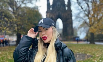 Mulher loira de cabelos longos posa ao ar livre usando casaco preto e boné dos Yankees; ao fundo, estrutura gótica alta cercada por árvores com folhas outonais.