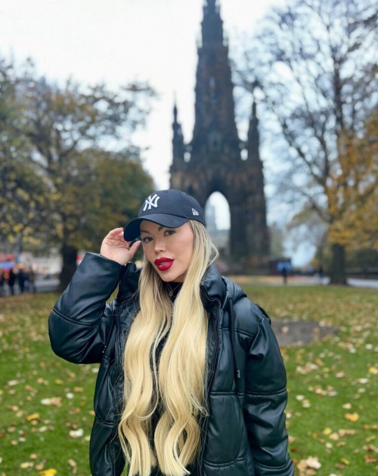 Mulher loira de cabelos longos posa ao ar livre usando casaco preto e boné dos Yankees; ao fundo, estrutura gótica alta cercada por árvores com folhas outonais.