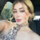 Mulher jovem com cabelo loiro preso em coque, usando blusa e lenço com estampa animal print, posando para selfie dentro de um carro. Ela tem olhos claros, maquiagem suave e expressão serena. Ao fundo, é possível ver vegetação e parte de uma construção externa pela janela.