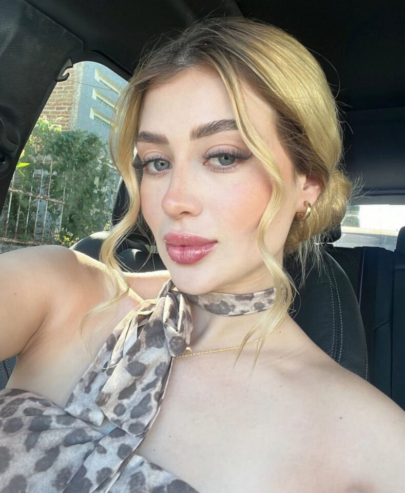 Mulher jovem com cabelo loiro preso em coque, usando blusa e lenço com estampa animal print, posando para selfie dentro de um carro. Ela tem olhos claros, maquiagem suave e expressão serena. Ao fundo, é possível ver vegetação e parte de uma construção externa pela janela.