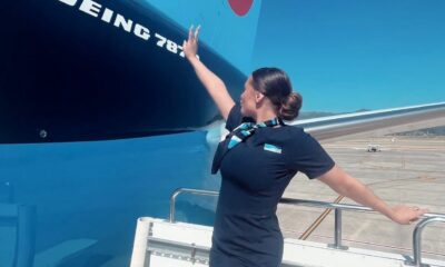 Comissária de bordo posa ao lado de um avião Boeing 787 em pista de aeroporto, vestindo uniforme preto com lenço azul e olhando para a fuselagem da aeronave